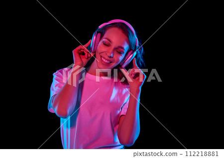 Cheerful, positive, smiling young woman in white t-shirt listening to music in headphones and dancing against black studio background in neon light Cheerful, positive, smiling young woman in white t-shirt listening to music in headphones and dancing against black studio background in neon light 112218881