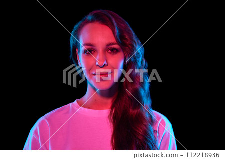 Portrait of beautiful, elegant young woman with curly hair posing with smile against black studio background in neon light Portrait of beautiful, elegant young woman with curly hair posing with smile against black studio background in neon light 112218936