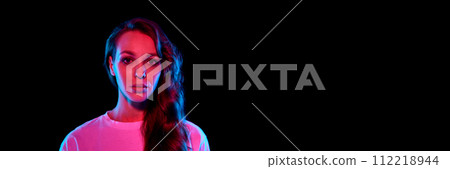 Portrait of beautiful young woman with brunette curly hair looking at camera against black studio background in neon light 112218944