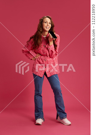 Beautiful young woman in casual clothes talking on mobile phone with smile, recording voice message against pink studio background Beautiful young woman in casual clothes talking on mobile phone with smile, recording voice message against pink studio background 112218950