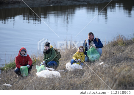 Little girl among eco activists picking up litter, along riverbank in nature, environmental pollution, eco activism and plogging concept. 112218965