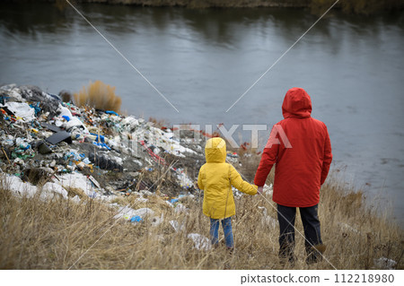 Little girl with father standing on river shore looking at piles of waste, debris on riverbank. Water and environmental pollution, eco activism concept. 112218980