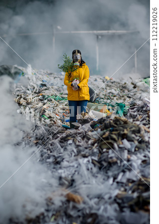 Activist with gas mask and plant standing on landfill, large pile of waste, environmental concept and eco activism. 112219026