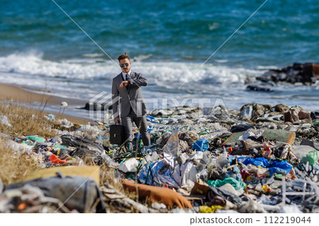 Businessman looking at watch, checking time standing on pile of waste on beach. Consumerism versus pollution concept. Corporate social responsiblity in business. Businessman looking at watch, checking time standing on pile of waste on beach. Consumerism versus pollution concept. Corporate social responsiblity in business. 112219044