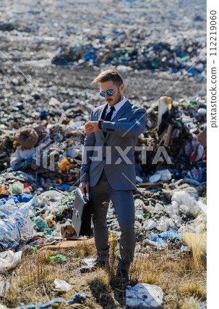 Businessman looking at watch, checking time standing on landfill, large pile of waste. Consumerism versus pollution concept. Corporate social responsiblity in business. 112219060