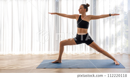Yoga woman in black sportswear standing in warrior pose on the mat in big room 112220118