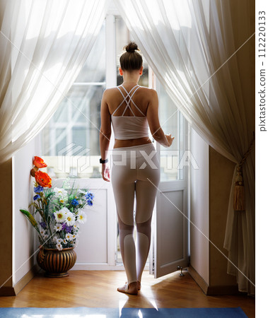 Young slim woman in white sportswear standing near open balcony in the morning 112220133