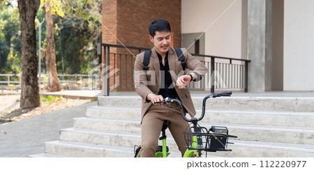 Asian businessman in a suit is riding a bicycle on the city streets for his morning commute to work. Eco transportation concept, sustainable lifestyle concept Asian businessman in a suit is riding a bicycle on the city streets for his morning commute to work. Eco transportation concept, sustainable lifestyle concept 112220977