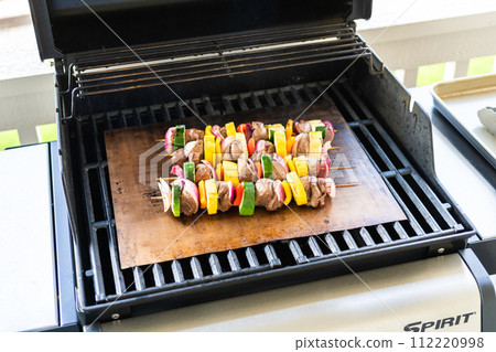 Skewered beef and fresh veggies sizzle on a copper grill mat over a gas outdoor grill. 112220998