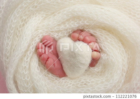 The tiny foot of a newborn baby. Soft feet of a new born in a white wool blanket. Close up of toes, heels and feet of a newborn. Knitted white heart in the legs of a baby. Macro photography The tiny foot of a newborn baby. Soft feet of a new born in a white wool blanket. Close up of toes, heels and feet of a newborn. Knitted white heart in the legs of a baby. Macro photography 112221076
