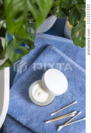 Opened cream jar on blue towels near basin and green monstera in bath, lid mockup 112221313