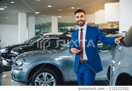 Confident auto dealer in modern car showroom, wearing sharp blue suit, presenting new car model, showcasing vehicle features and options to potential buyers 112222582