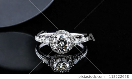 Image captures mesmerizing sparkle of diamond-studded white gold ring, elegance amplified by dark, reflective backdrop that accentuates every detail of this luxurious piece of jewelry. 112222658