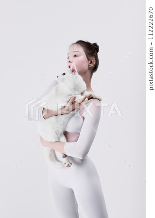 Wellness brand campaigns promoting the peaceful coexistence with pets. Young woman in white clothes gently interacts with a white cat against white studio background 112222670
