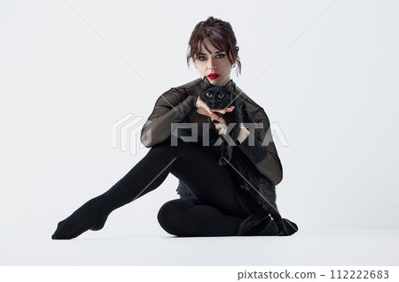 Connection with animals. Passionate young woman with smoky eyes and red lips makeup, black clothes, posing with black cat against white studio background 112222683
