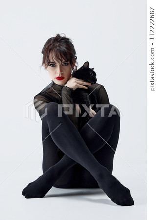 Beautiful young woman with smoky eyes and red lips makeup, in black outfit, sitting and holding adorable black cat against white studio background 112222687