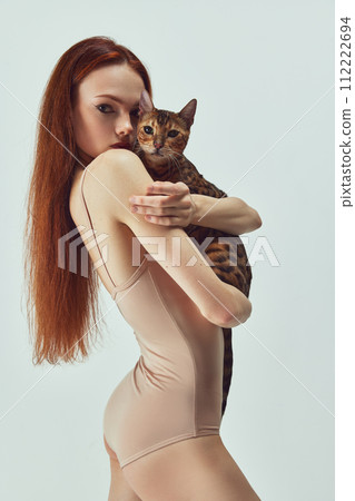 Bond between human and pet. Beautiful young redhead woman with Bengal cat against white studio background 112222694