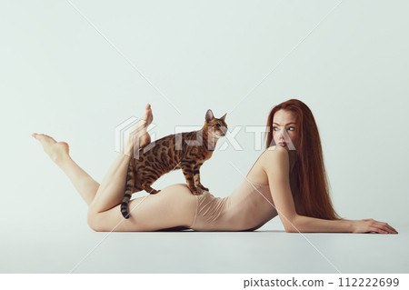 Passion and tenderness. Young redhead beautiful girl with long hair, slim body lying on floor with purebred Bengal cat against white studio background Passion and tenderness. Young redhead beautiful girl with long hair, slim body lying on floor with purebred Bengal cat against white studio background 112222699