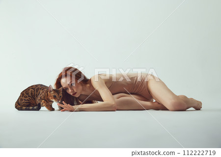 Beautiful, young, elegant redhead woman in beige bodysuit lying with purebred Bengal at against white studio background 112222719