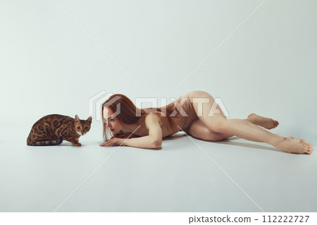 Beautiful, young, elegant redhead woman in beige bodysuit lying with purebred Bengal at against white studio background 112222727