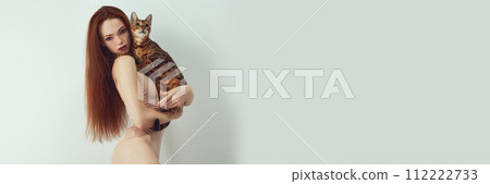 Femininity and self-identity. Elegant young redhead woman with slim body holding purebred Bengal cat against white studio background 112222733