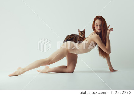 Femininity and self-identity. Passionate, beautiful young redhead woman with slim body with purebred Bengal cat lying on back against white studio background Femininity and self-identity. Passionate, beautiful young redhead woman with slim body with purebred Bengal cat lying on back against white studio background 112222750