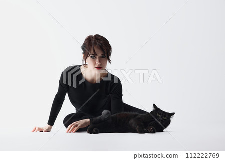 Bond between human and pet. Beautiful young woman in black clothes sitting on floor, posing with black cat against white studio background 112222769