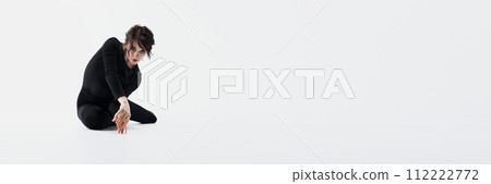 Elegant passionate young woman in black clothes, with dark eyes makeup sitting on floor with emotive face against white studio background 112222772