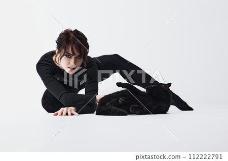 Connection with animals. Passionate young woman with smoky eyes makeup, deep look, black clothes, posing with black cat against white studio background Connection with animals. Passionate young woman with smoky eyes makeup, deep look, black clothes, posing with black cat against white studio background 112222791