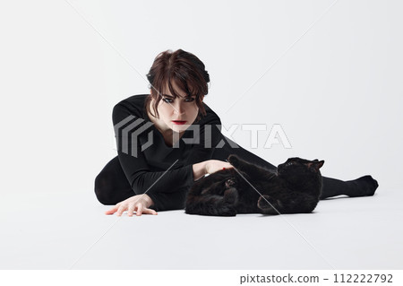 Connection with animals. Passionate young woman with smoky eyes makeup, black clothes, posing with black cat against white studio background 112222792