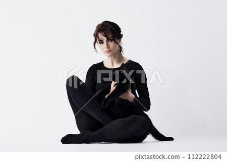 Beautiful young woman with smoky eyes makeup, in black outfit, sitting and holding adorable black cat against white studio background Beautiful young woman with smoky eyes makeup, in black outfit, sitting and holding adorable black cat against white studio background 112222804