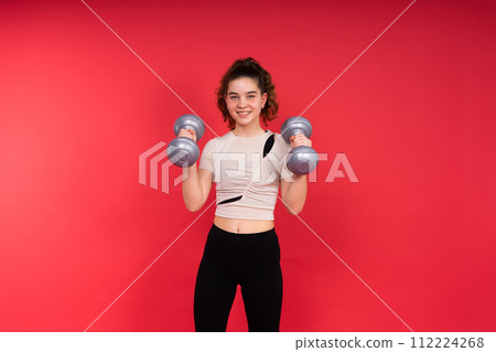 Teenage sportive girl exercises with dumbbells to develop muscles isolated on red background. 112224268