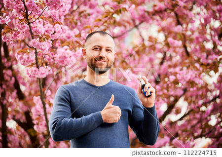 Man allergic using medical nasal drops, suffering from seasonal allergy at spring in blossoming garden. Portrait of smiling man showing thumbs up near blooming tree outdoors. Spring allergy concept. 112224715