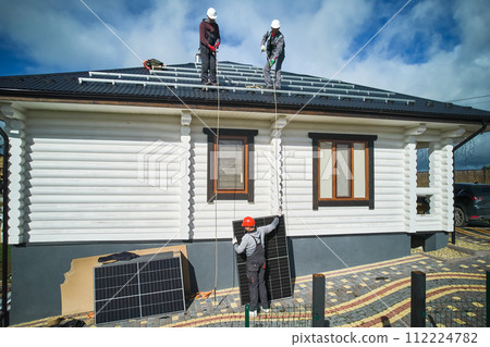 Workers lifting up solar panel on a roof of house. Three male workers in helmets and work overalls carrying solar cell. Concept of alternative energy resources. Workers lifting up solar panel on a roof of house. Three male workers in helmets and work overalls carrying solar cell. Concept of alternative energy resources. 112224782