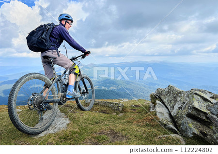 Cyclist man riding electric bike outdoors. Male tourist resting on the top of hill, enjoying beautiful mountain landscape, wearing helmet and backpack. Concept of active leisure. Cyclist man riding electric bike outdoors. Male tourist resting on the top of hill, enjoying beautiful mountain landscape, wearing helmet and backpack. Concept of active leisure. 112224802