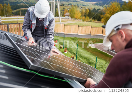 Technicians building photovoltaic solar module station on roof of house. Men engineers in helmets installing solar panel system outdoors. Concept of alternative and renewable energy. Technicians building photovoltaic solar module station on roof of house. Men engineers in helmets installing solar panel system outdoors. Concept of alternative and renewable energy. 112224805