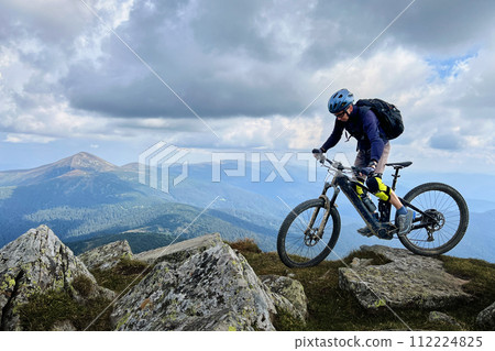 Cyclist man riding electric mountain bike outdoors. Male tourist biking along grassy trail in the mountains, wearing helmet and backpack. Concept of sport, active leisure and nature. 112224825