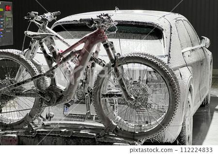 Electric vehicle and bicycles at self-service car wash during nighttime, covered in foam, indicating that it is in the middle of a wash cycle. Bicycles mounted on a car rack at the back. Electric vehicle and bicycles at self-service car wash during nighttime, covered in foam, indicating that it is in the middle of a wash cycle. Bicycles mounted on a car rack at the back. 112224833