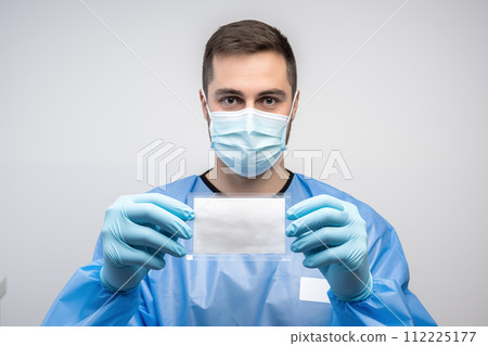 Portrait of a young male doctor in protective mask and gloves holding blank card 112225177