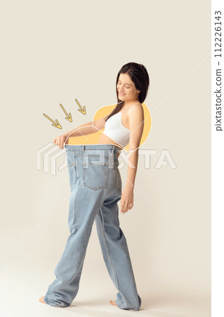 Smiling woman showcasing her weight loss by wearing oversized jeans against grey studio background. Smiling woman showcasing her weight loss by wearing oversized jeans against grey studio background. 112226143
