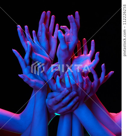 Group of hands reaching upwards showing uplifting and supportive in multicolored neon lighting against black studio background. 112226258