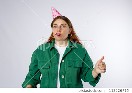 Young happy woman in birthday party hat with playing pipe showing thumbs up gesture. Emotional, young face. Human emotions, facial expression and holiday concept. Young happy woman in birthday party hat with playing pipe showing thumbs up gesture. Emotional, young face. Human emotions, facial expression and holiday concept. 112226715