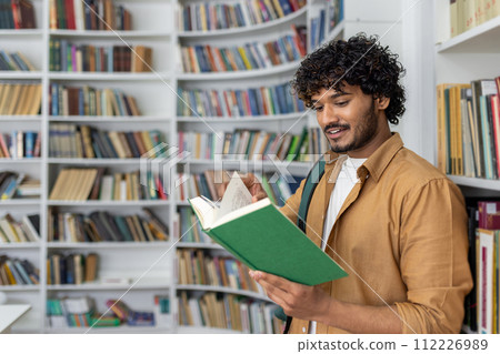 An adult male stands in a well-lit library, immersed in reading a green book. Bookshelves filled with various books surround him, suggesting a quiet atmosphere for study and literature exploration. 112226989