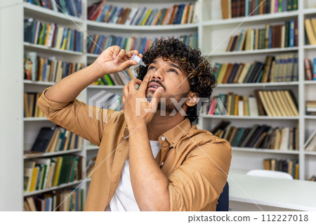 A young adult male is captured applying eye drops while standing in a cozy home library. The image reflects self-care and health management against a backdrop of bookshelves filled with books. A young adult male is captured applying eye drops while standing in a cozy home library. The image reflects self-care and health management against a backdrop of bookshelves filled with books. 112227018
