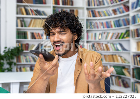 A young man with curly hair appears exasperated while having a heated conversation on his smartphone, standing against a backdrop of bookshelves in a library setting. A young man with curly hair appears exasperated while having a heated conversation on his smartphone, standing against a backdrop of bookshelves in a library setting. 112227023