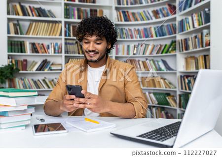 A young, casually dressed professional engages with a smartphone while seated at a desk covered with books, a laptop, and a tablet, surrounded by shelves filled with books in modern library. A young, casually dressed professional engages with a smartphone while seated at a desk covered with books, a laptop, and a tablet, surrounded by shelves filled with books in modern library. 112227027