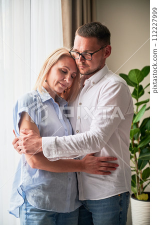 Happy couple hugging on light background at home. Pretty woman smiling and hugging her husband with closed eyes. Sweet couple in casual clothes. 112227999