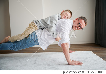 Energetic father and his little son training at home. Kid climbed dad's back while he doing plank. Young family enjoying activity games at home. Energetic father and his little son training at home. Kid climbed dad's back while he doing plank. Young family enjoying activity games at home. 112228003
