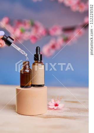 Glass bottle with pipette and serum on a podium and branches of cherry blossoms Glass bottle with pipette and serum on a podium and branches of cherry blossoms 112228582