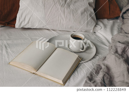 Cup of coffee, open books on white bed sheet. Breakfast in bed concept. Blurred muslin, linen pillows, blanket. Moody cozy Scandinavian bedroom. Top view, no people. Lifestyle banner. Cup of coffee, open books on white bed sheet. Breakfast in bed concept. Blurred muslin, linen pillows, blanket. Moody cozy Scandinavian bedroom. Top view, no people. Lifestyle banner. 112228849